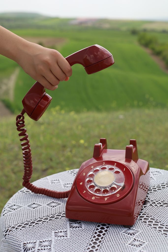 A hand lifting a red rotary phone receiver, set against a lush green field background.
